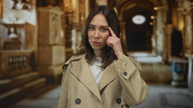 Bej ceketli genç İspanyol kadın bir kilise ortamında düşüncelere dalmış bir şekilde duruyor..