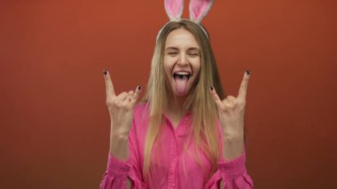 Paskalya tavşanı kulağı takan ve iki eliyle kaya jesti yapan bir kadın, izole edilmiş kırmızı bir arka plana yaslanmış, heyecan ve neşe ifade ediyor..