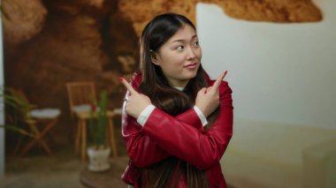 Kapalı bir yerde kırmızı ceketli çapraz kollarıyla poz veren bir kadın, rahat bir otel arka planına karşı kurulmuş, yön duygusunu ve karar verme mekanizmasını aktarıyor..