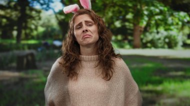 Parkta genç bir kadın üzgün bir ifadeyle tavşan kulağı takıyor, açık hava ortamında yemyeşil ile çevrili, doğada oyunculuk ve duygunun bir karışımını yakalıyor..