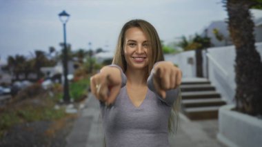 Dışarıdaki kamerayı gösteren kadın, güveni olan sokak arkasını gösteriyor. Gri gömlekli, gülen, öğleden sonra aydınlık şehir manzaralı..