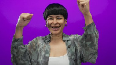 Kolları parlak mor arka planda gülümseyen, neşe ve heyecan ifade eden, şık gömlek ve küpeler giyen bir kadın..