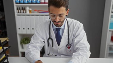 Klinikteki genç İspanyol erkek doktor steteskop ve beyaz önlük giyip modern tıbbi çalışma ortamında profesyonel odaklı belgeleri inceliyor..