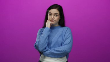 Parlak pembe bir arka plana karşı duran, mavi bir kazak giyen, kumral saçlı, çapraz kolları ve düşünceli bakışlarıyla tefekkür eden bir kadın..