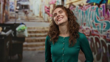 Grafiti dolu şehir sokaklarında gülümseyen bir kadın, yeşil bir kazak giymiş, gözleri kapalı neşeli ve tasasız bir yüz ifadesi sergiliyor..