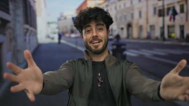 Sakallı genç adam dışarıda, güneşli sokakta gülümsüyor. Mutluluk ve kendine güveni taşıyor. Canlı şehir ortamında bulanık binalar ve arka planda insanlar var..