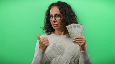 Kafası karışmış bir ifadeyle Dirham banknotları tutan ve sağlam yeşil bir zemine karşı baş parmak kaldıran bir kadın belirsizlik ve mali karar alma duyusunu iletiyor..