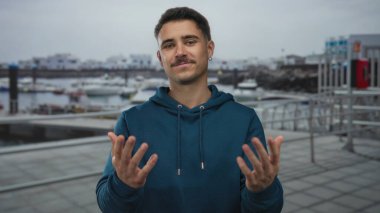 Mavi eşofmanlı genç İspanyol adam deniz kenarında hareketli hareketler yapar, bulutlu bir günde arka planda tekneler ve deniz kenarındaki binalarla hareketli bir limanda durur.