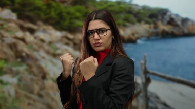 Sahilde güven içinde poz veren, gözlük takan, manzaralı bir okyanus zeminine karşı güç ve kararlılık sergileyen, zarafeti doğanın güzelliğiyle harmanlayan bir kadın..