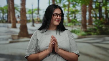 Uzun siyah saçlı, uzun boylu, gözlüklü ve gri gömlekli, palmiye ağaçları ve bulanık yeşilliklerle çevrili genç bir kadın..