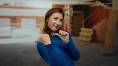 İnşaat alanında mavi bir kazakla gülümseyen bir kadın, başparmaklarıyla arkasını gösteriyor, ev dekorasyonunda neşe ve güven gösterisi yapıyor..