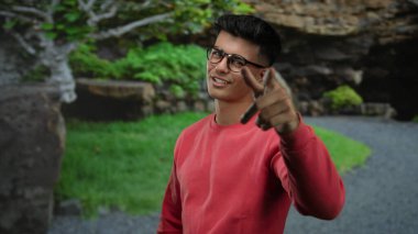 Gözlüklü ve kırmızı kazaklı genç adam, parlak bir günde yemyeşil bir parkta güvenle dikiliyor..