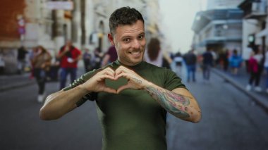 Dövmeleri olan genç bir adam şehir sokaklarında kalp şekli oluşturur. Arka planda bulanık yayalar şehir ortamında sevgi ve bağlantı hissi taşır.