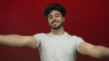 Kolları açık gülümseyen genç bir adam canlı kırmızı bir arka planda neşeli bir hoş geldin jesti sergiliyor sakallı ve günlük beyaz bir tişört giyiyor..