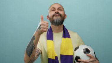 Sakallı, elinde futbol topu olan, mavi arka planda baş parmak gösteren, sarı mor eşarplı, güvenli bir şekilde poz veren beyaz bir adam..