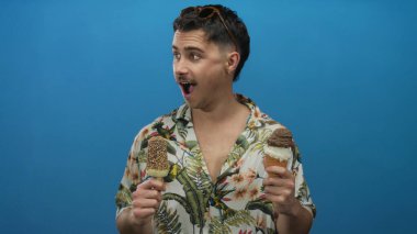 Tropikal tişörtlü genç adam, elinde parlak mavi bir arka planda iki dondurma tutarken, neşeli ve hareketli bir yaz turisti anını yakalıyor..