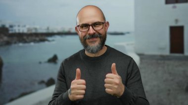 Deniz kenarındaki gezinti alanının önünde elleri dövmeli gülümseyen gözlüklü, süveterli bulanık plajlı ve arka planda binalar olan bir adam..