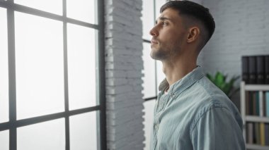 Sıradan gömlekli genç adam, modern iç mekanda, tuğla duvarlar ve raflarla geniş ofis penceresinden dikkatlice bakıyor, yansıtıcı ve profesyonel bir atmosfer yaratıyor..