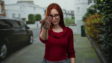 Kadın şehir sokaklarında jest yapıyor, gözlük takıyor ve kırmızı kazak giyiyor, kızıl saçlı duygularını ifade ediyor, şehir merkezinde iletişim kurmak için kamerayı işaret ediyor..