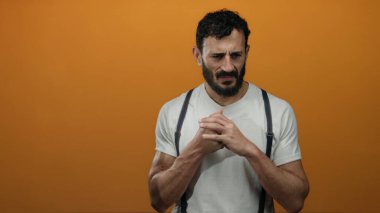 Sakallı İspanyol adam canlı turuncu arka planda duruyor, beyaz gömlek ve pantolon askısı giyiyor, çeşitli düşünceli ifadeler sergiliyor..