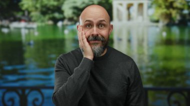 Parkta bir adam yüzüne sürpriz bir ifadeyle dokunuyor, sıradan bir süveter giyiyor, yemyeşil manzaraya ve gölün arka planına karşı duruyor..