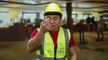 Otel lobisinde inşaat kıyafeti giyen bir adam kapalı alanda yoğun bir ortamda kararlı bir görünüm sergiliyor.