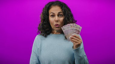 Mor arka planda Meksika pezosu tutan orta yaşlı bir kadın elinde banknotlarla şaşırmış görünüyor, izole bir ortamda zenginlik ve duyguyu ön plana çıkarıyor..