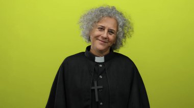 Gri saçlı, ruhban kıyafeti giyen ve haçlı kolyesi olan yaşlı bir kadın dingin tavırlı, sakin ve şefkatli bir tavırla hayat dolu sarı bir arkaplan karşısında duruyor..