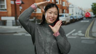 Açık havada gülümseyen bir kadın gri kazak giymiş, canlı poz veren genç Çinli bir kadın kentsel arka planda canlı enerji sergiliyor..