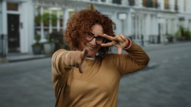 Şehir sokaklarında barış işareti yapan gözlüklü bir kadın ve açık havada neşeli bir ifade gösteren kahverengi bir kazak..