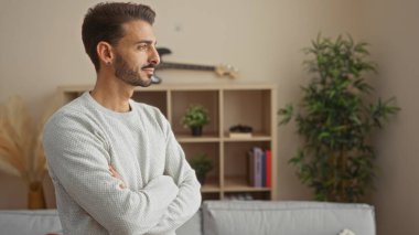 Şık oturma odasında kollarını kavuşturmuş genç bir adam, modern iç dekorasyonu bitkiler ve gitarla sergiliyor, rahat ve çağdaş bir hava veriyor..