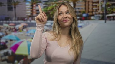 Sarışın kadın açık güneşli gökyüzünün altında kumlu sahili işaret ediyor; merak uyandıran keşif merakı.