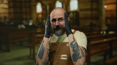 Dövmeli ve eldivenli kel bir adam kilisede yüzünü örtüyor. Duygularını iletiyor ve gündüz vakti kutsal kapalı bir ortama odaklanıyor..