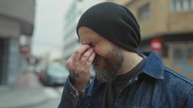 Şehir caddesinde kel sakallı bir adam. Yüzünde elleri, kot ceketi ve beresi var..