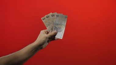 Finansal kavramları ve parasal temaları sembolize eden, çarpıcı bir kırmızı arkaplan karşısında izole edilmiş Türk lirası banknotları tutan bir adamın eli.