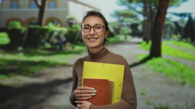 Güneş ışığı parkında gülümseyen dosyalar taşıyan genç bir kadın eğlence yaratıcılığını sergiliyor ve çevresi yemyeşil ve açık mavi gökyüzü ile çevrili açık havada öğreniyor..