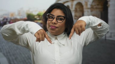 Roma Kolezyumu 'nda gözlüklü genç bir kadın, aşağı işaret ediyor, beyaz gömlek giyiyor, kendine güveni tam bir İspanyol, büyük beden modası, tarihi mimariye karşı..