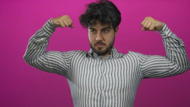 Sakallı ve çizgili gömlekli genç adam canlı pembe arka plana karşı kaslarını esnetiyor..