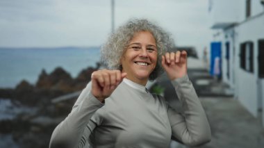 Gri saçlı yaşlı bir kadın deniz kenarında manzaralı bir arka planda neşeyle dans ediyor, açık havada mutluluk ve özgürlüğün özünü yakalıyor..