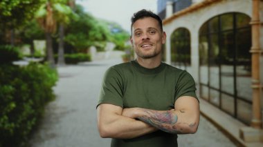 Yeşil gömlekli gülümseyen adam ağaçlarla ve binalarla dolu güneşli bir caddede kollarını kavuşturarak rahat ve kendine güvenen bir atmosfer yaratıyor.