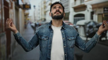 Sakallı İspanyol adam düşünceli bir şekilde şehir caddesinde duruyor, kot giyiyor, duacı hareketlerle duygularını ifade ediyor, ruhani bir doğa deneyimi sunuyor..