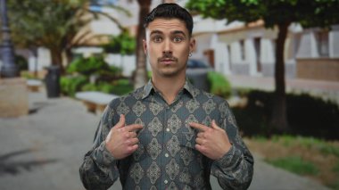 Genç İspanyol bir adam açık hava parkı ortamında sürpriz bir jest yapıyor, arka planda ağaçlar ve banklar olan desenli bir gömlek giyiyor, samimi bir kendini tanıma anını yakalıyor..