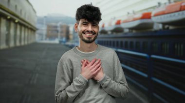 Deniz kıyısında teknelerle gülümseyen, gri bir kazak giyen ve neşeli bir ifade sergileyen genç adam, liman çevresinin sükunetini ve çekiciliğini yakalıyor..