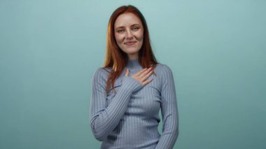 Mavi arka planda, göğsünde bir elle duran, gri bir kazak giyen, kızıl saçlı, yetişkin ve güzel bir yüz ifadesi olan, sakin ve sakin bir atmosferi olan bir kadın..