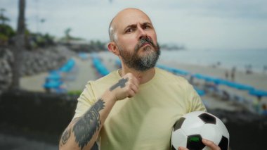 Kel sakallı adam deniz kıyısındaki arka planda bulanık futbol topuyla tehditkar hareketler yapıyor..