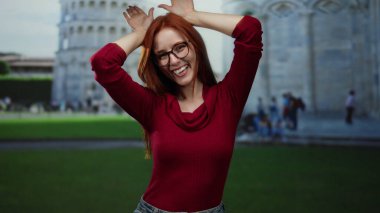 Kırmızı kazaklı kızıl kadın, güneşli bir günde pisa kulesinin önünde elleri kulaklarıyla neşeli bir şekilde poz veriyor. Eğlenceli bir seyahat anı yakalıyor..