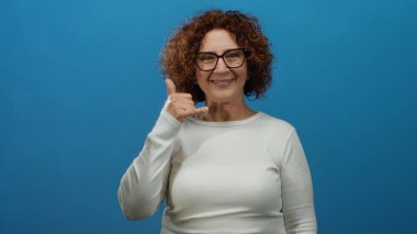 Gülümseyen bir kadın, canlı, mavi bir arka plana karşı bir telefon jesti yaparken orta yaşlı İspanyol sıcaklığı ve bağlantı sergiliyor..