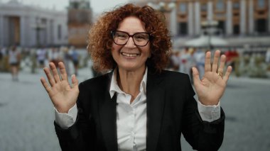 Orta yaşlı, siyah takım elbiseli, gülümseyen İspanyol kadın arka planda hareketli şehir hayatı olan Aziz Peter Meydanı, Vatikan 'da duruyor..