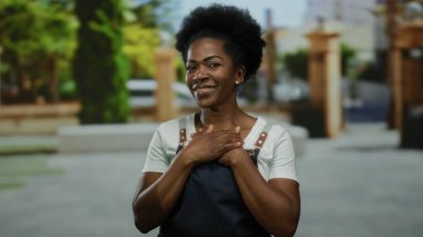 Kadın açık havada göğsünde ellerle gülümsüyor, önlük giyiyor, bir şehir caddesi düzeninde, kentin ortasında bir memnuniyet ve minnettarlık duygusu somutlaştırıyor..