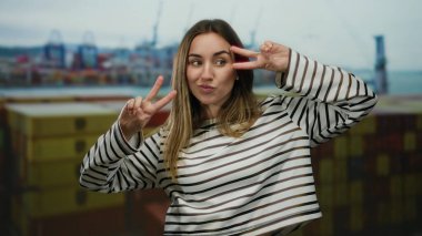 Limanda çizgili süveter giyen, arka planda bulutlu bir gökyüzünün altında nakliye konteynerleri ve vinçleri olan bir kadın gülümsüyor..